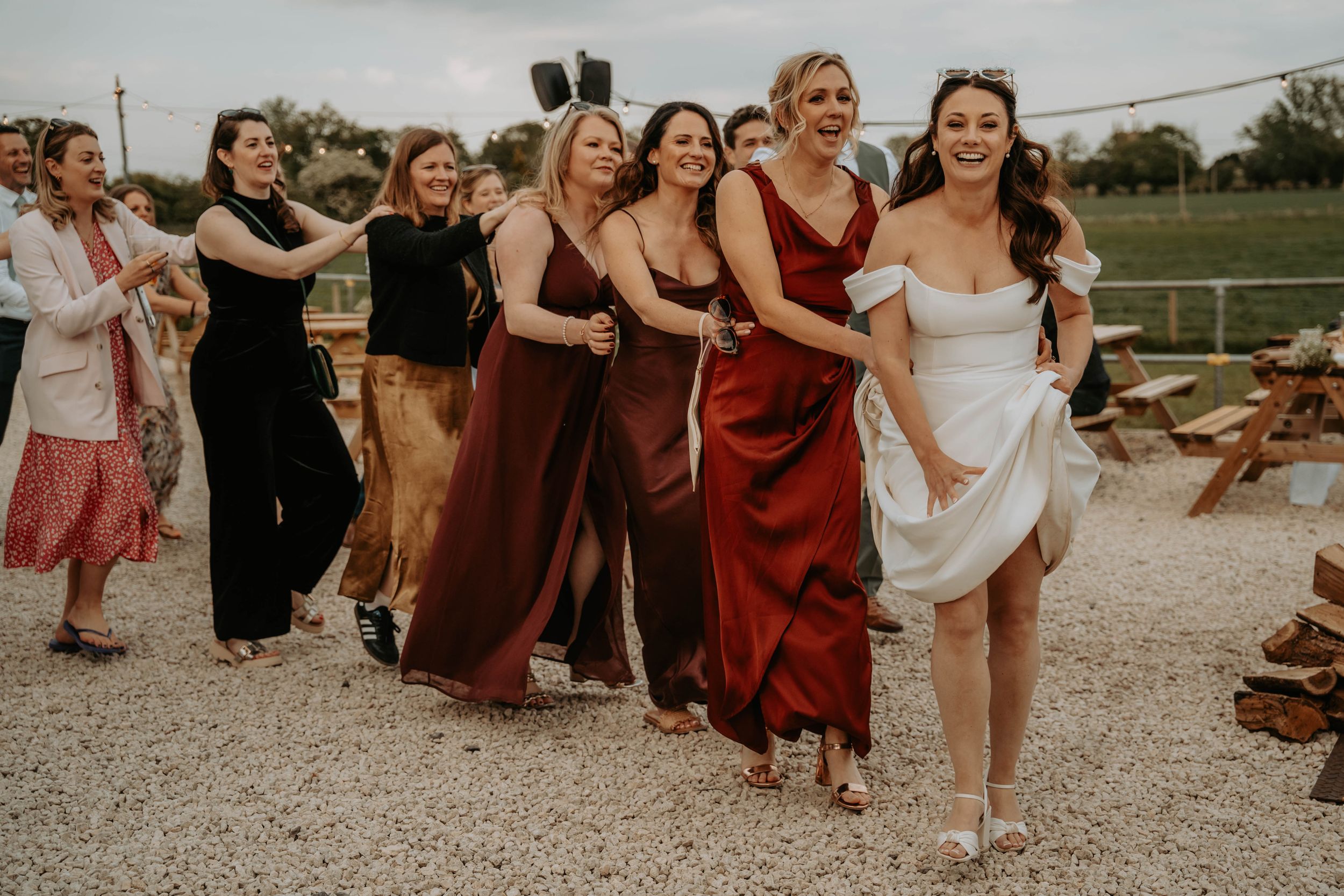 Bride in white dress laughing and running with bridesmaids in burgundy and black gowns at outdoor wedding reception.