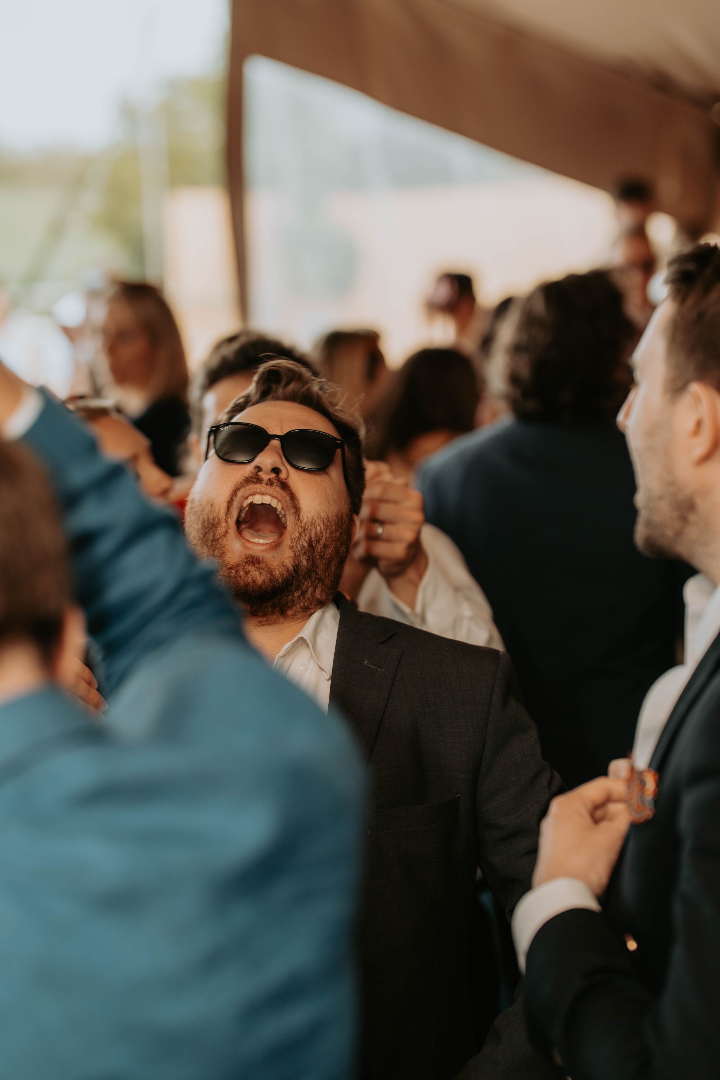 A bearded man in sunglasses and suit laughs joyfully at a crowded wedding reception party.