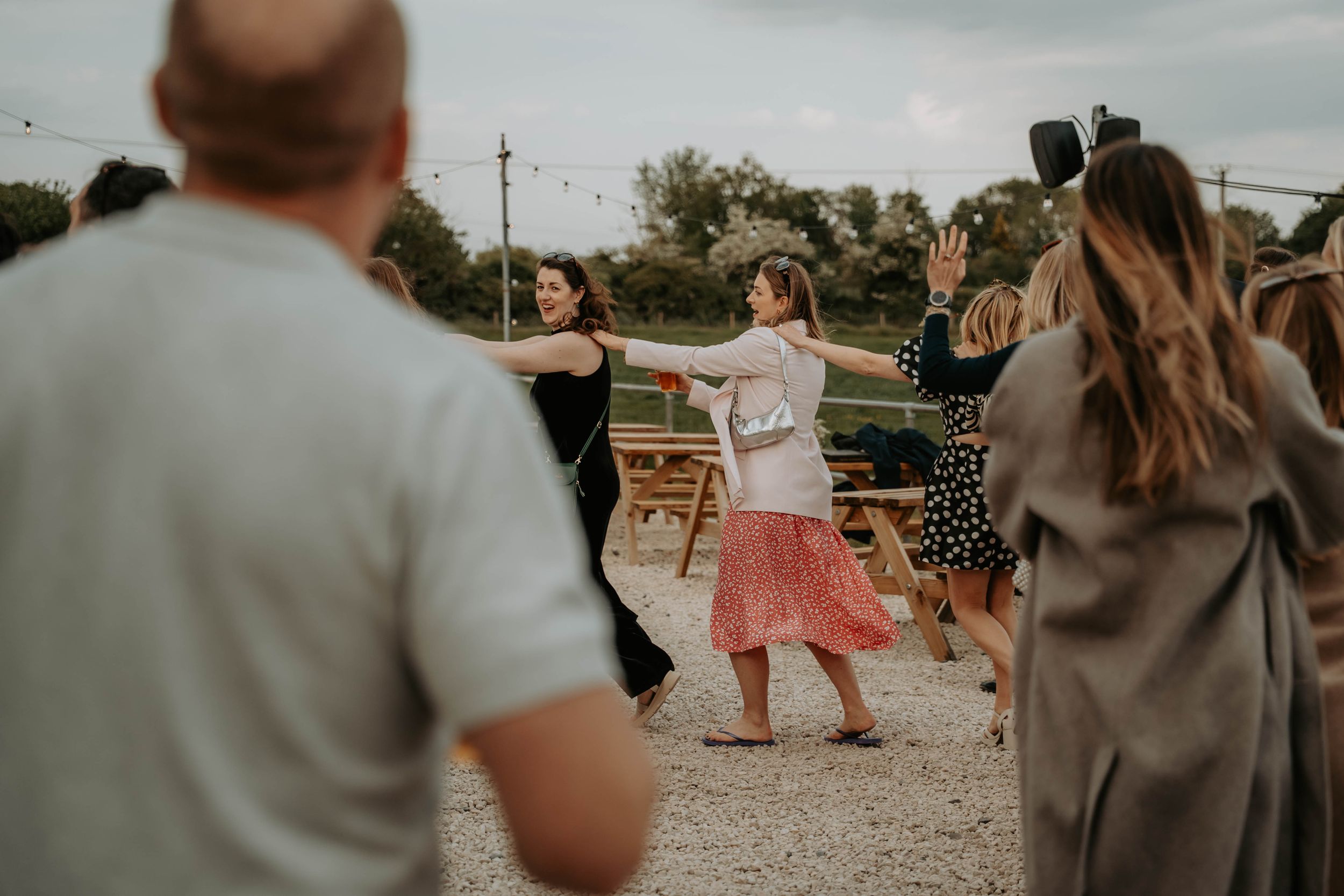 People dancing outdoors at a casual gathering, with a girl in a pink skirt spreading her arms wide on a gravel path.