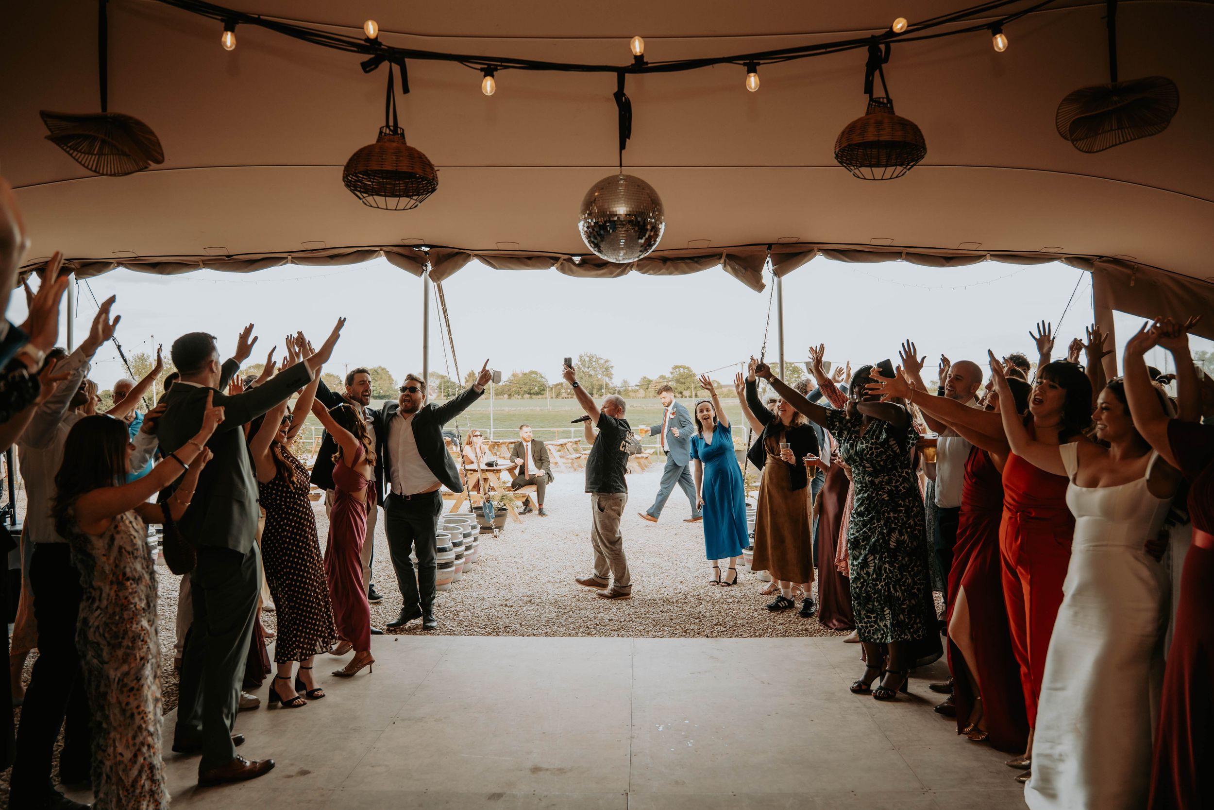 Wedding guests raise hands as couple walks through tent aisle under string lights and disco ball.