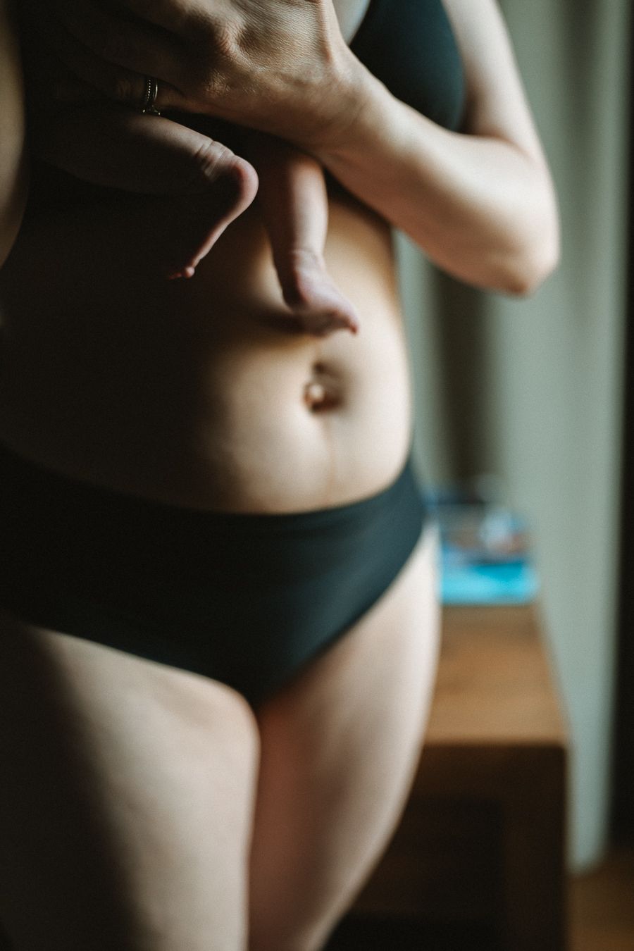 Woman in dark bikini set posing indoors, showcasing natural body curves in soft natural lighting.