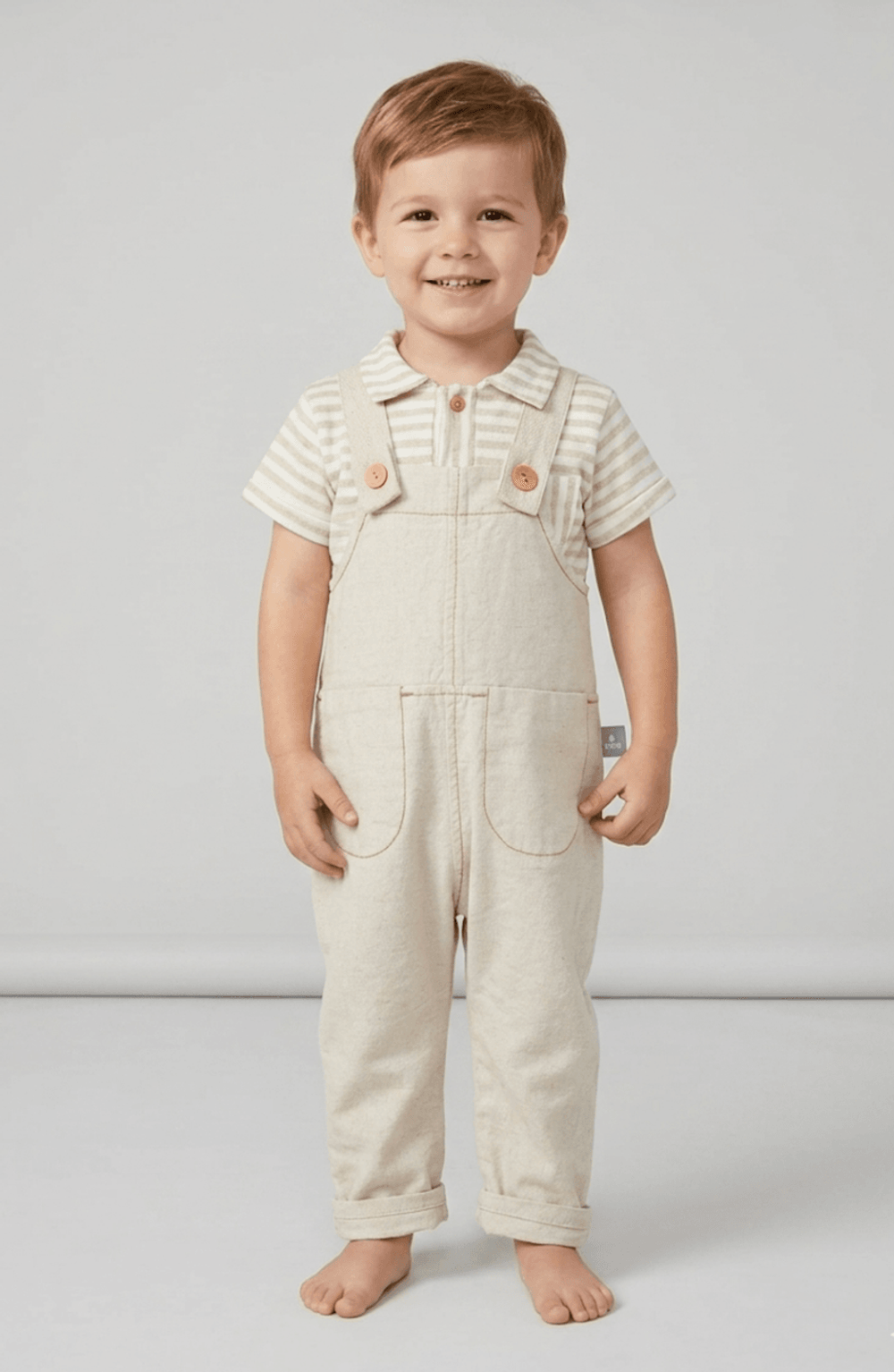 Toddler boy wearing beige striped polo shirt and neutral linen overalls, smiling in a clean studio setting.