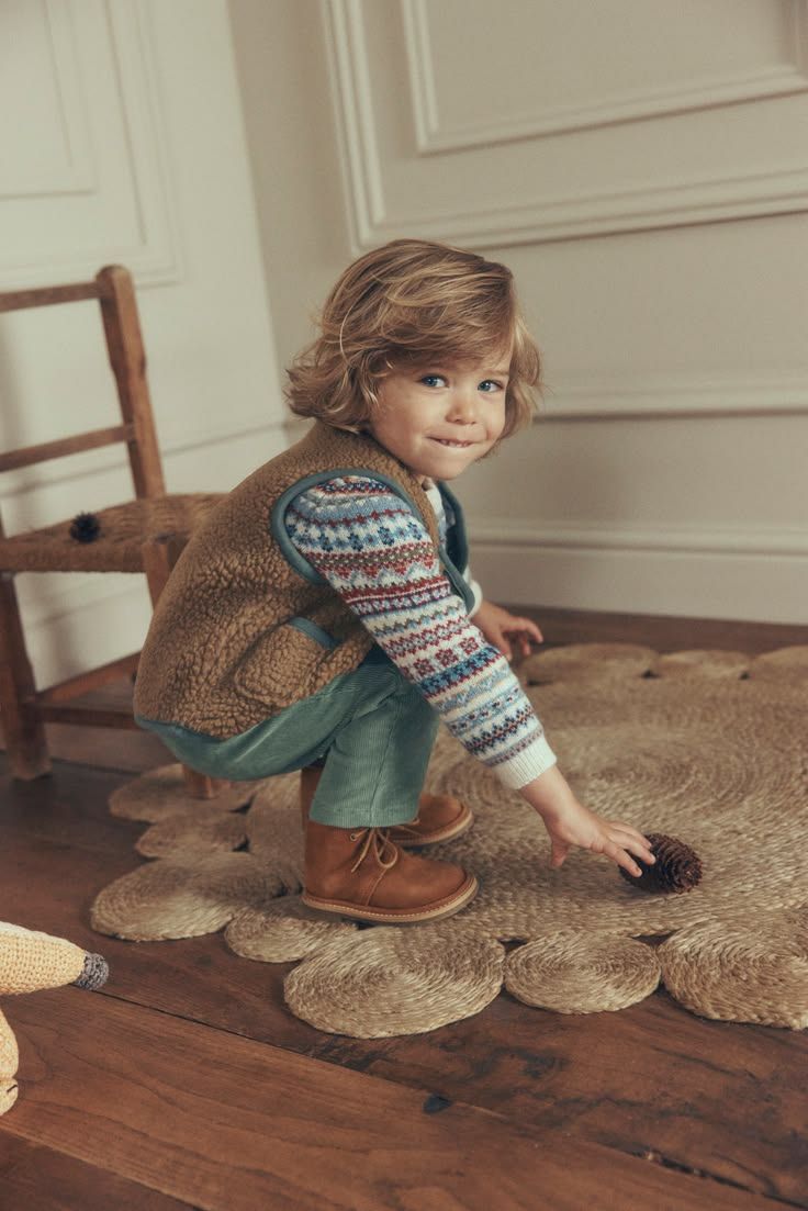 Toddler boy in fair isle sweater, green pants and brown boots playing on a jute rug indoors.