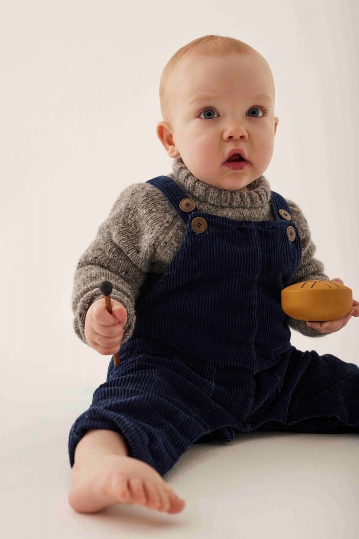 Baby boy in navy corduroy overalls and grey knit turtleneck sweater sits barefoot holding a wooden toy.