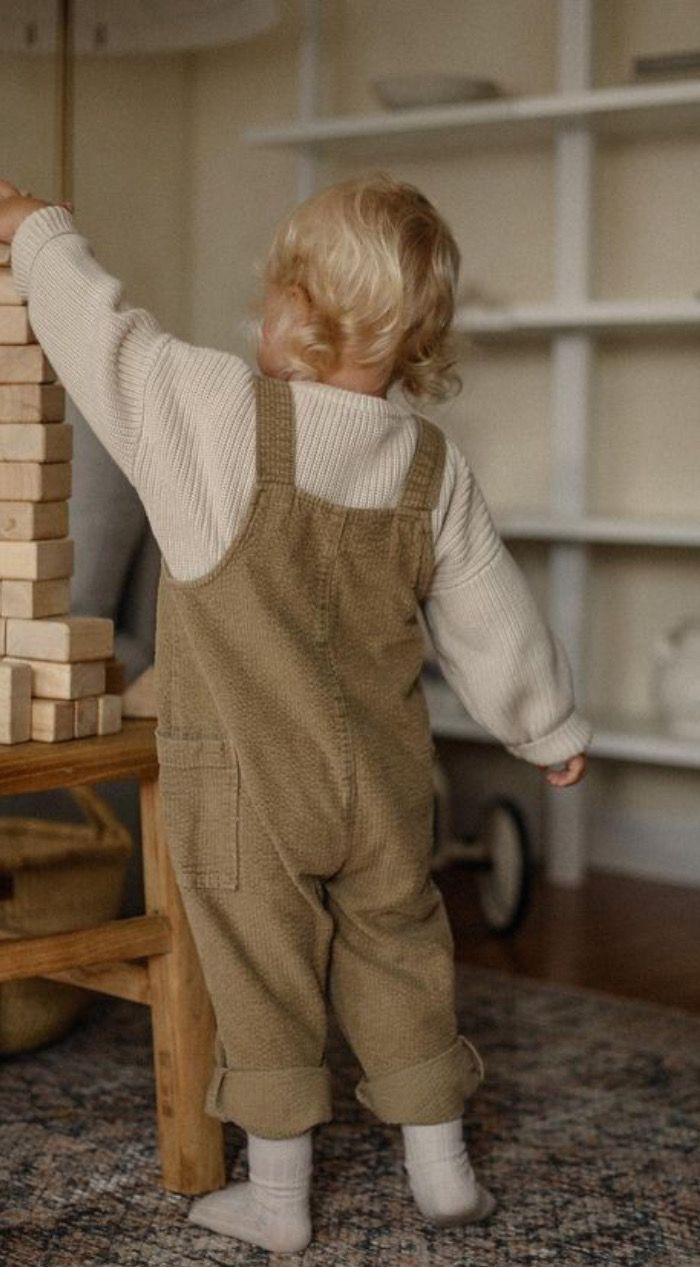 Toddler in olive corduroy overalls and cream sweater plays with wooden blocks, viewed from behind.