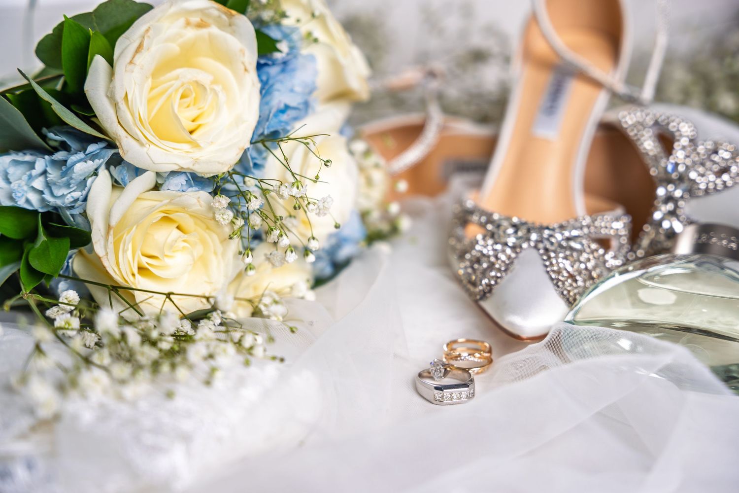 Elegant wedding flat lay with white roses, blue hydrangeas, diamond heels, and gold rings on white fabric.