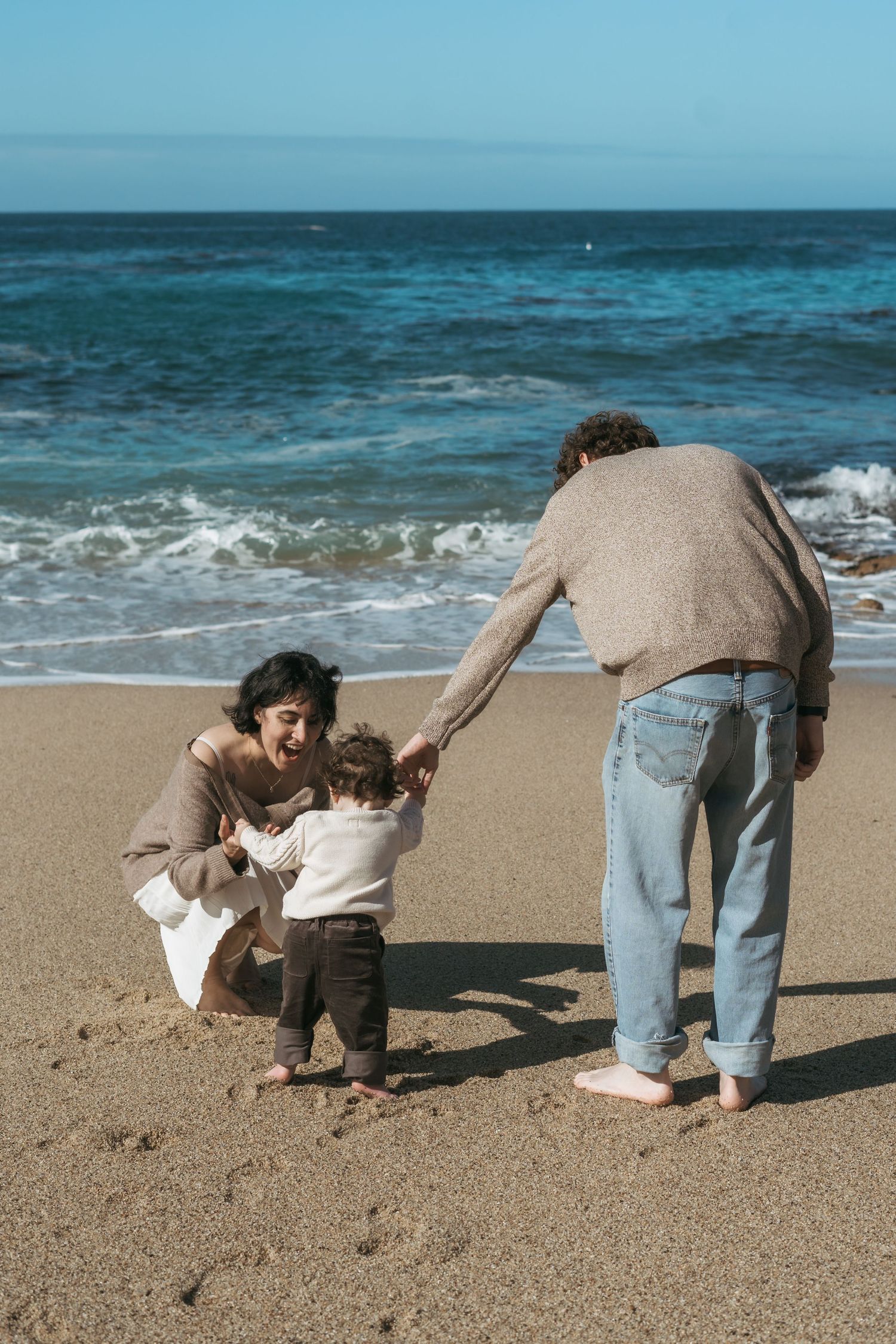 Family with toddler enjoying a sunny beach day, parents helping baby walk on wet sand by ocean waves.