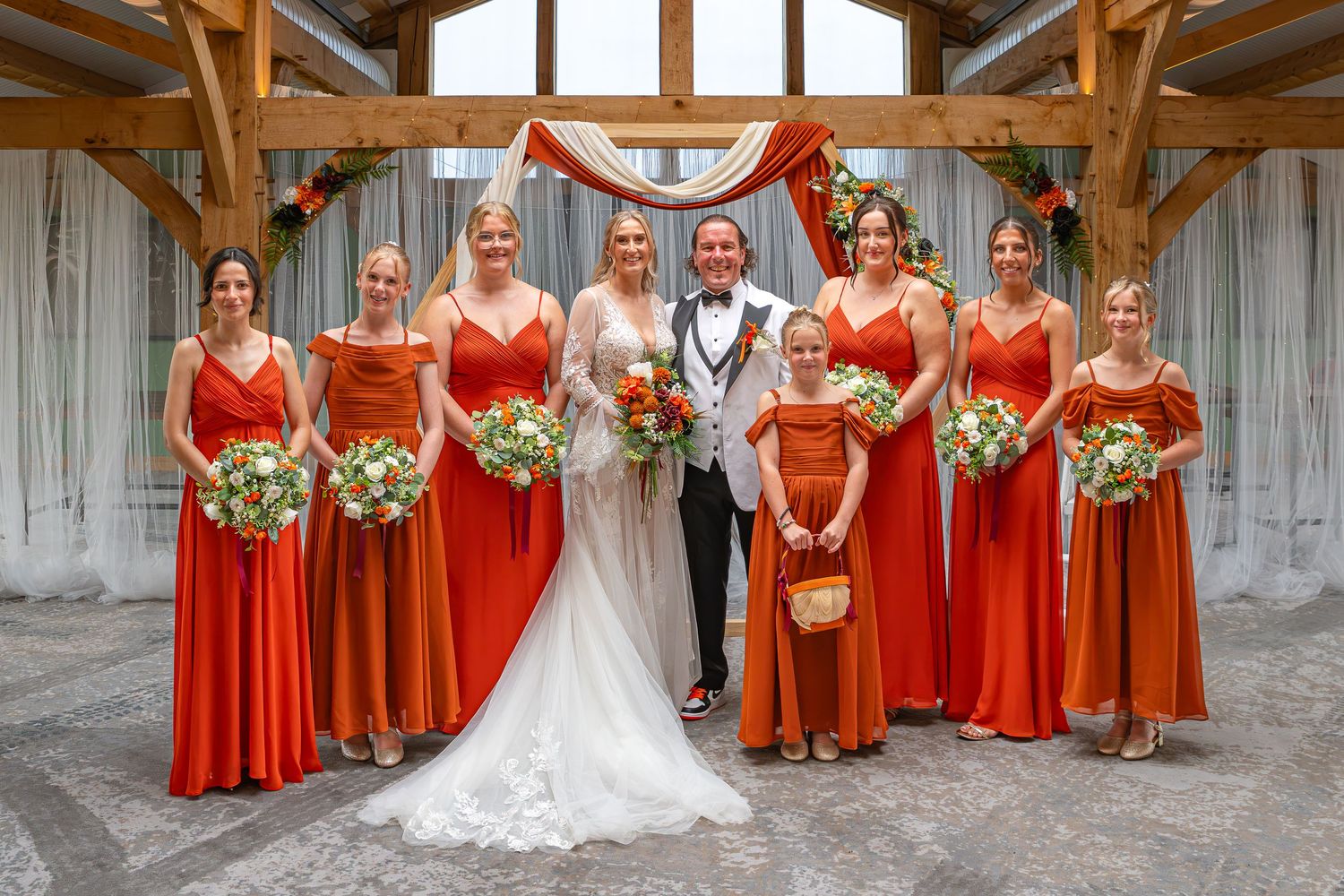 Wedding party poses in rustic venue, bride and groom flanked by bridesmaids in orange dresses holding green bouquets.