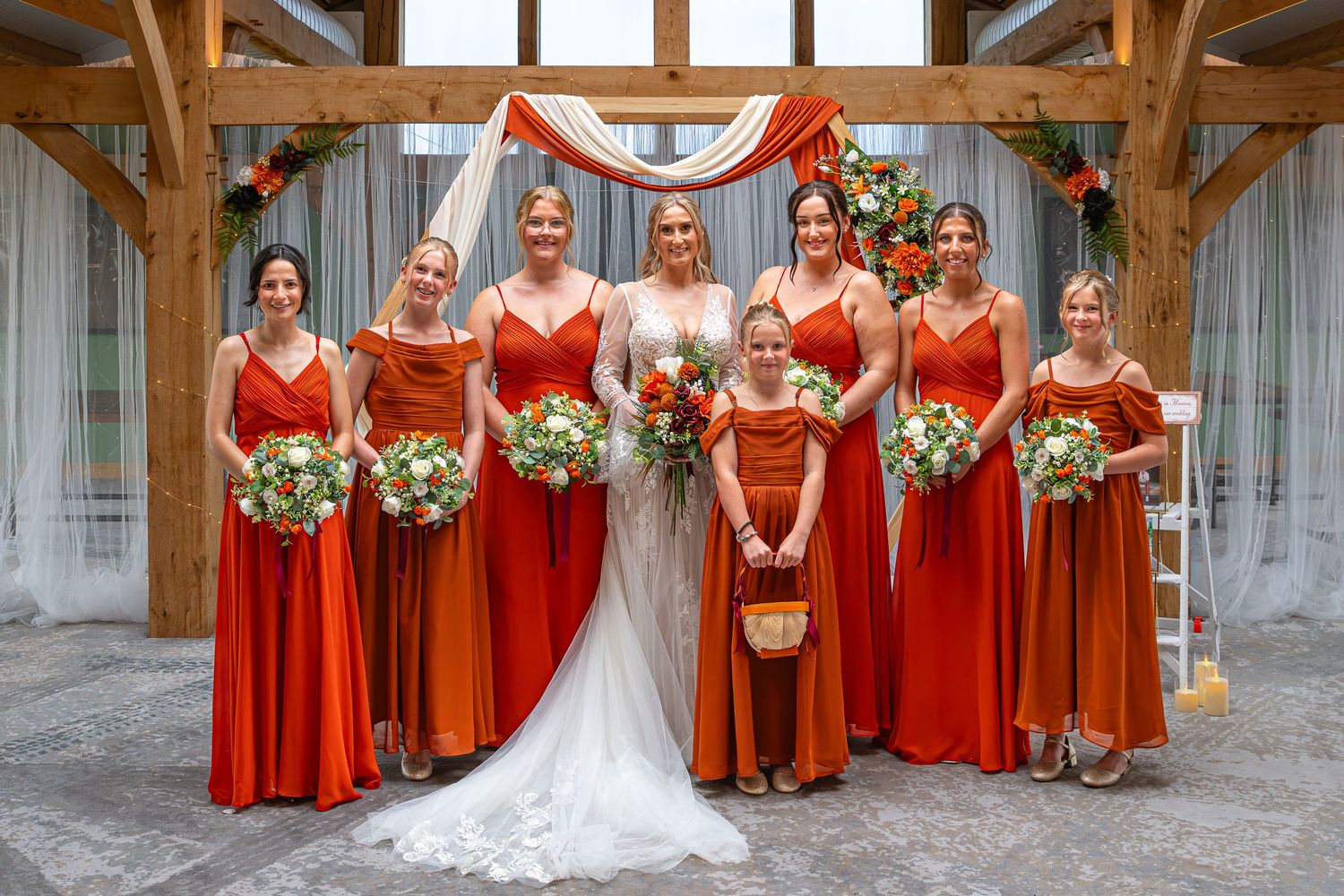 Bride with bridesmaids in orange dresses and flower girl holding bouquets at rustic barn wedding ceremony.Yorkshire, Sheffield, Lincolnshire
