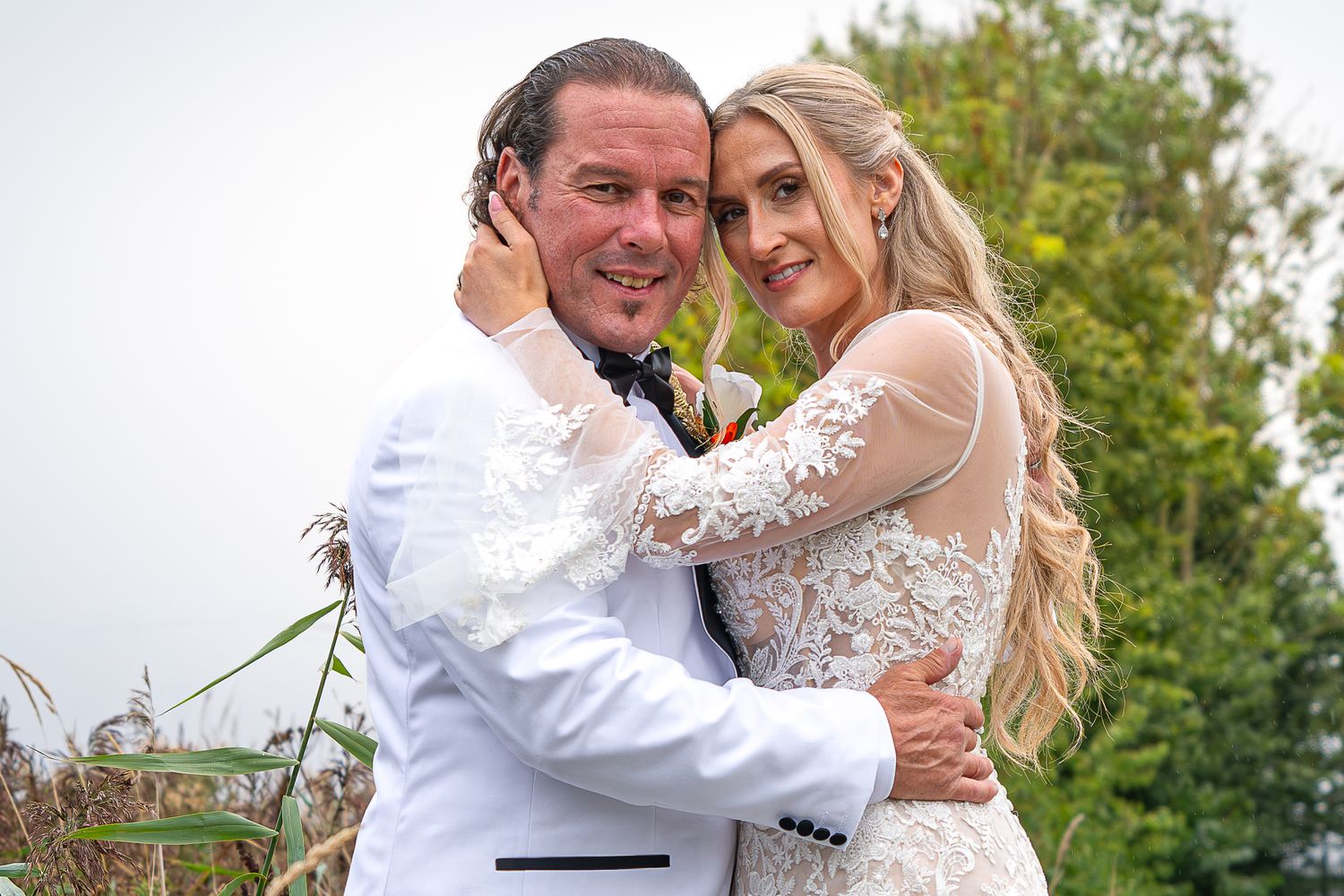 A smiling couple in wedding attire embrace outdoors, the groom in a white suit and bride in a lace gown.Yorkshire, Sheffield, Lincolnshire,
