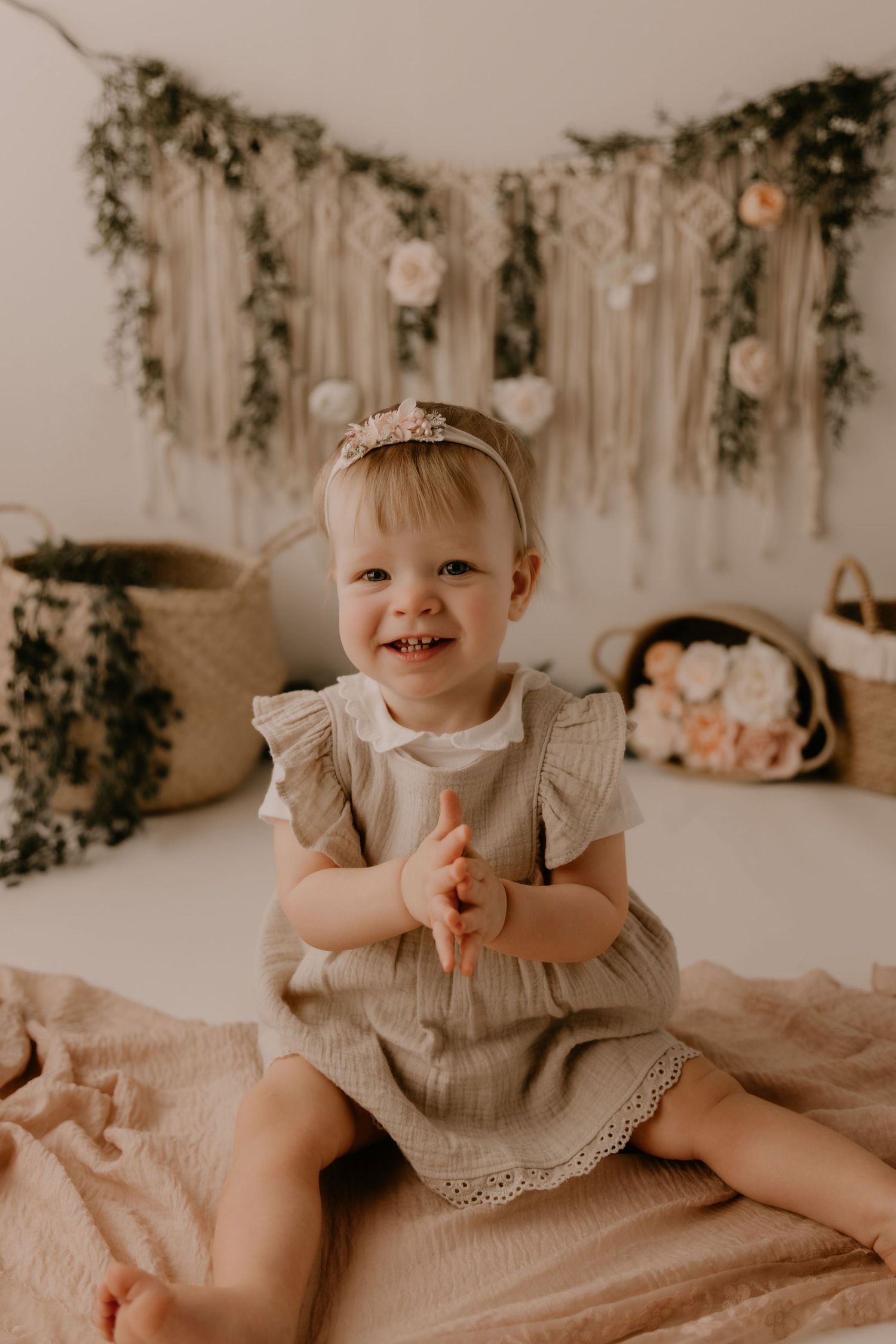 Smiling toddler girl in linen dress sits on pink blanket in boho studio with macrame wall hanging and floral baskets.