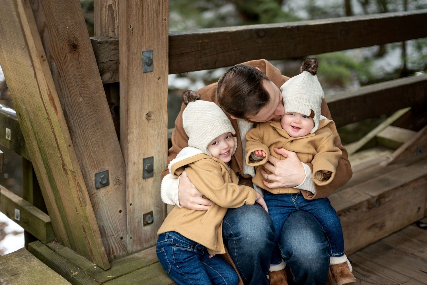 Parent hugging two young children in matching cream hats and tan coats on a wooden deck in winter.