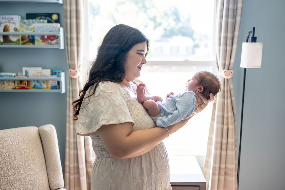 A young mother in a white dress tenderly holds her newborn baby in a softly lit nursery room.