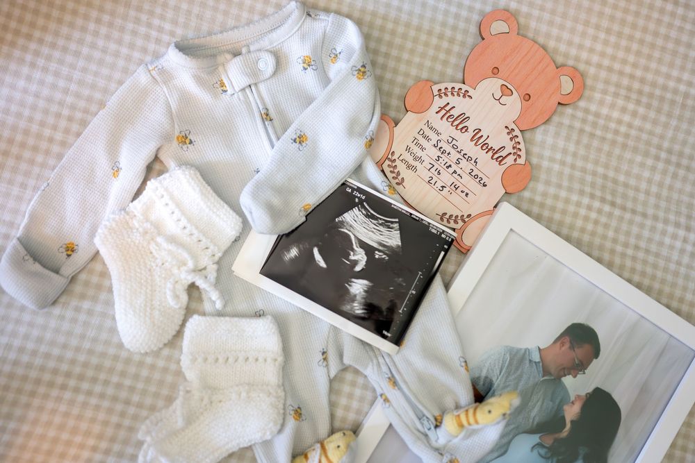 Baby shower flat lay with white onesie, socks, ultrasound photo, birth announcement card, and couple photo on checkered background.