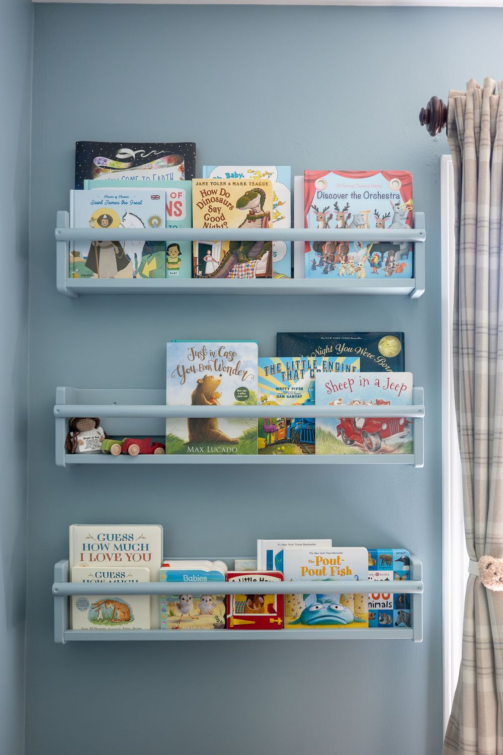 Three white wall-mounted ledge bookshelves filled with colorful childrens picture books in a blue nursery.