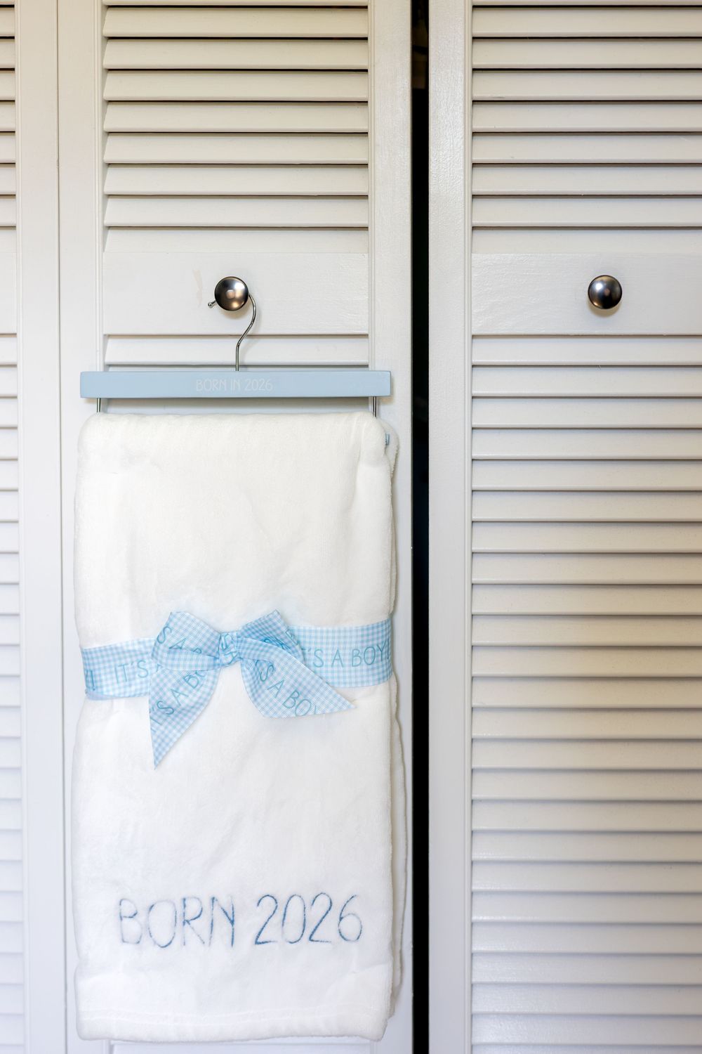 White blanket with blue bow and Born 2026 text hanging on a hanger against white louvered closet doors.