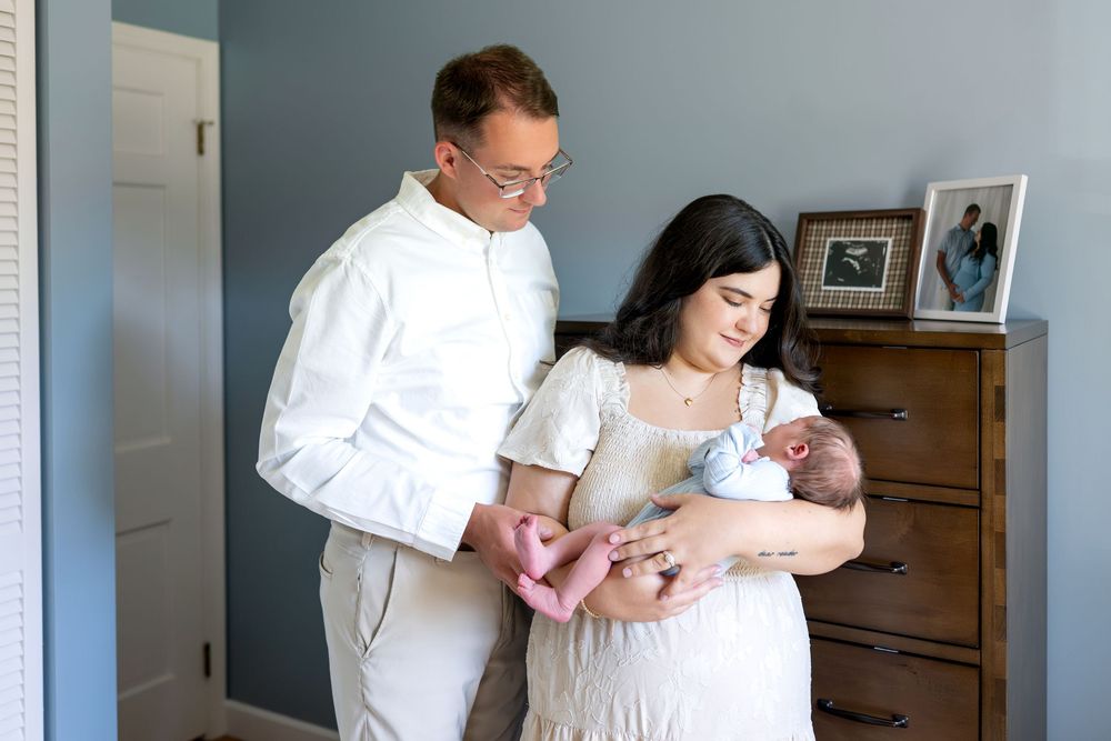 New parents in white outfits lovingly gaze at their newborn baby in a softly lit nursery room.