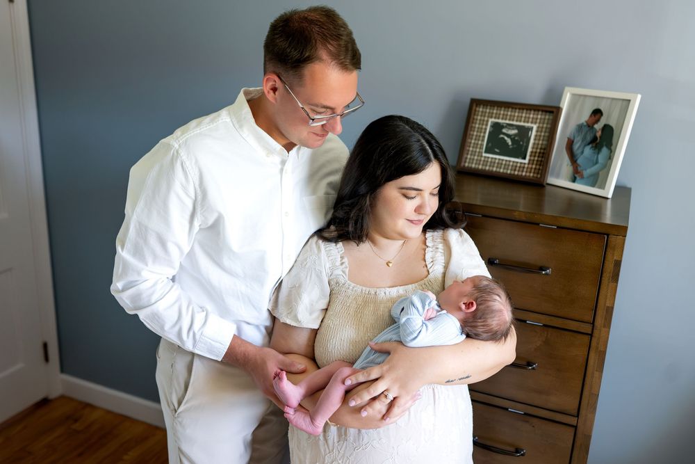 New parents tenderly hold their sleeping newborn baby in a cozy nursery with a wooden dresser.