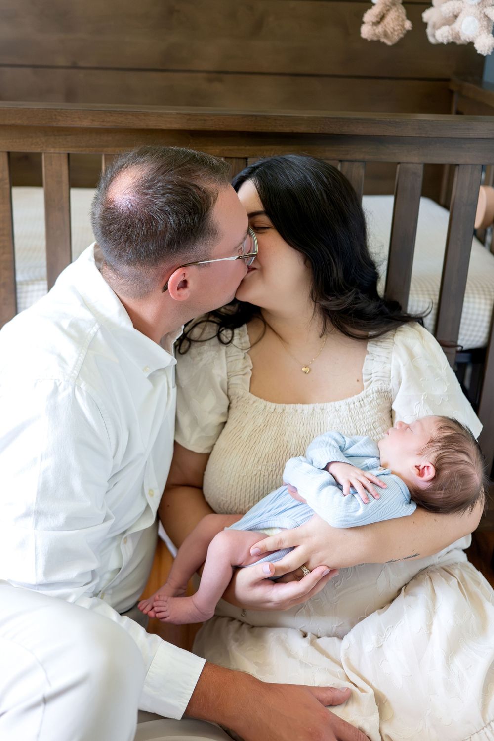 New parents kissing while cradling their sleeping newborn in a blue onesie near a wooden crib.