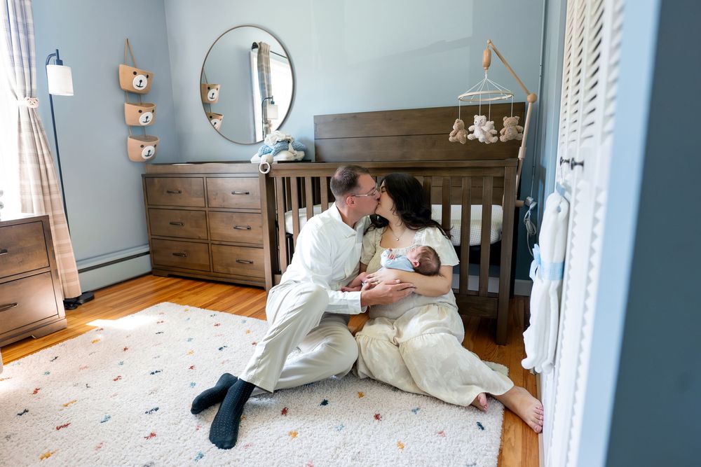 New parents sit on nursery floor holding newborn baby, sharing a tender kiss beside a wooden crib.
