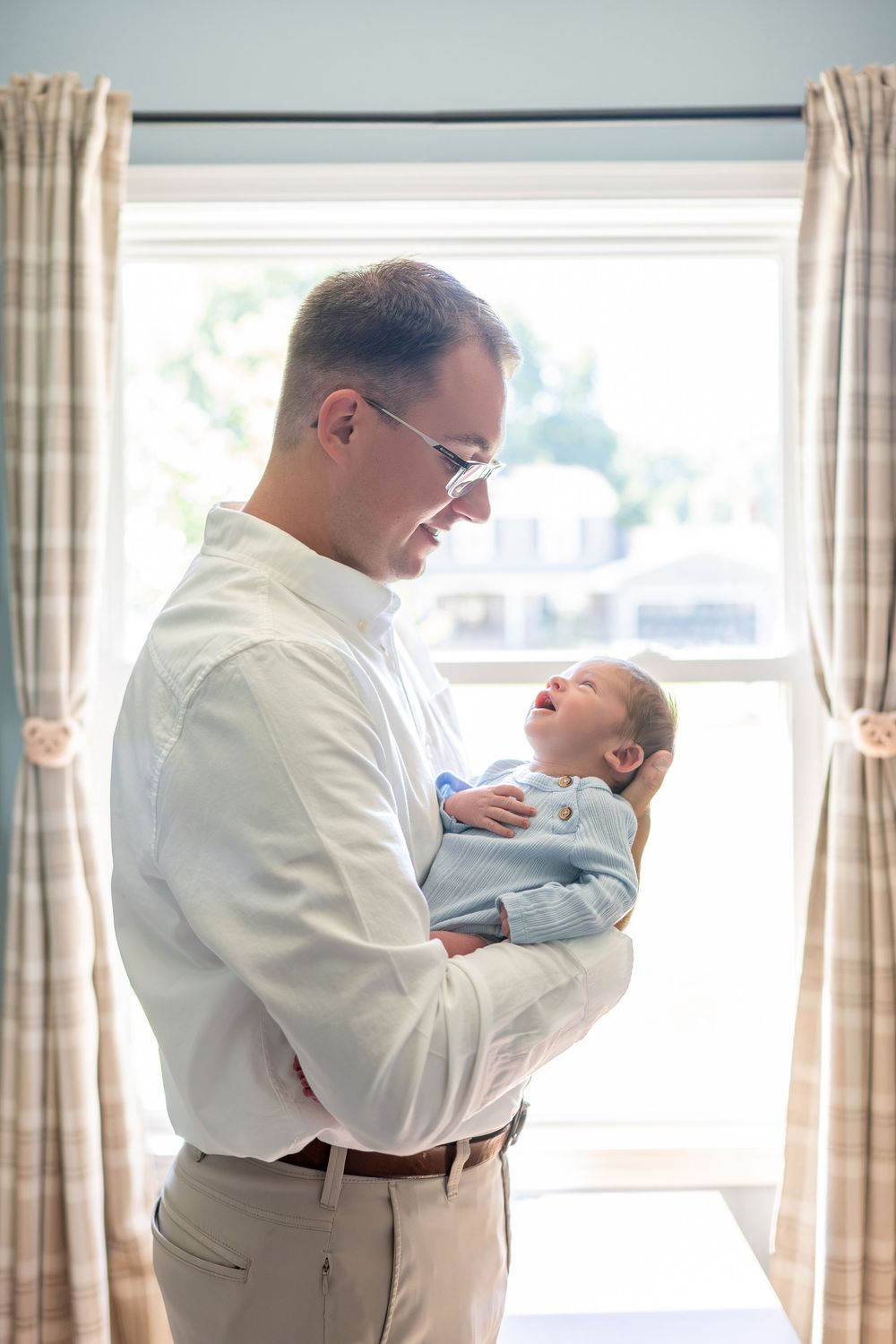 Father smiling down at his newborn baby in a blue outfit, standing by a bright window with plaid curtains.