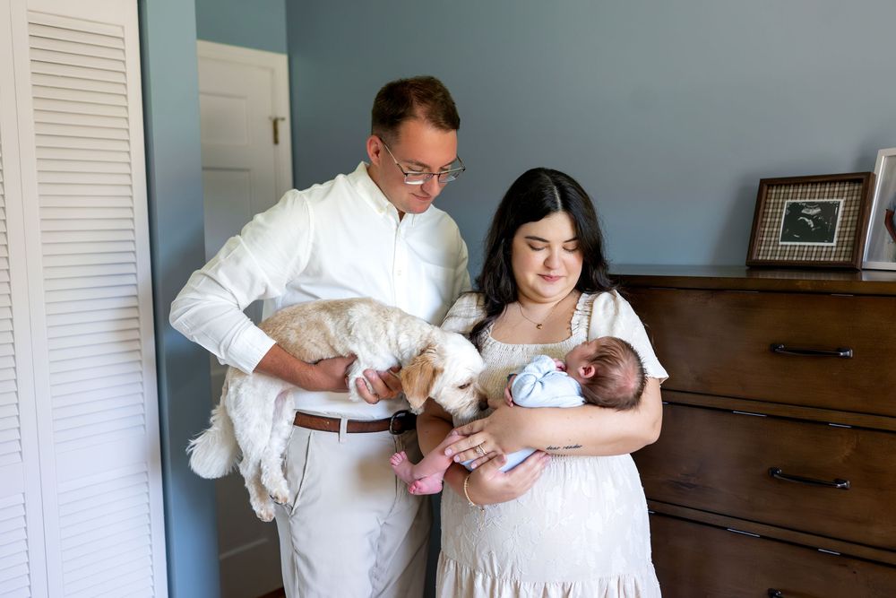 New parents holding newborn baby and fluffy puppy in cozy nursery bedroom.
