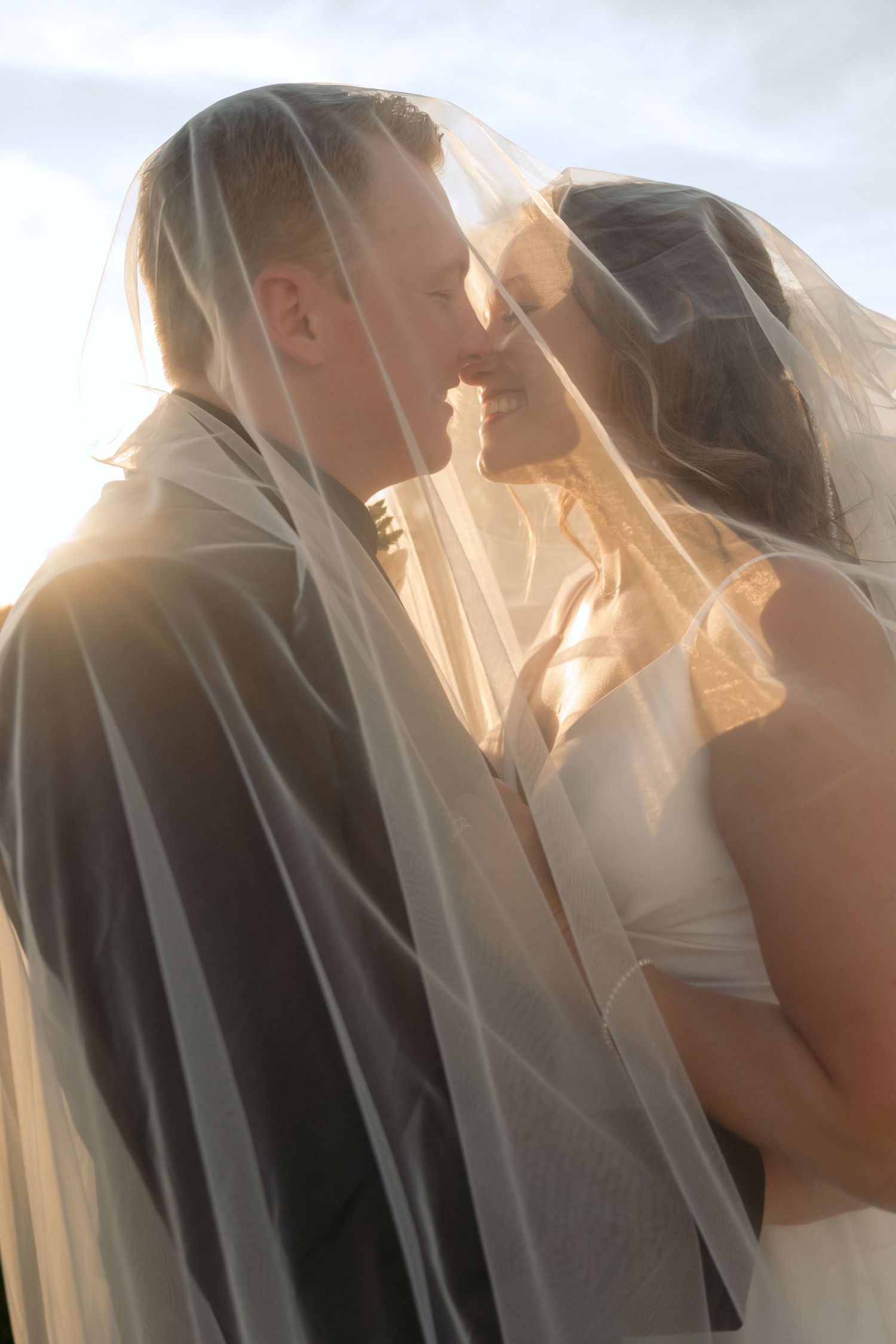 Bride and groom smile and go in for a kiss while covered in the bride's veil during sunset portraits at Abella Farm in Chisago City, MN.