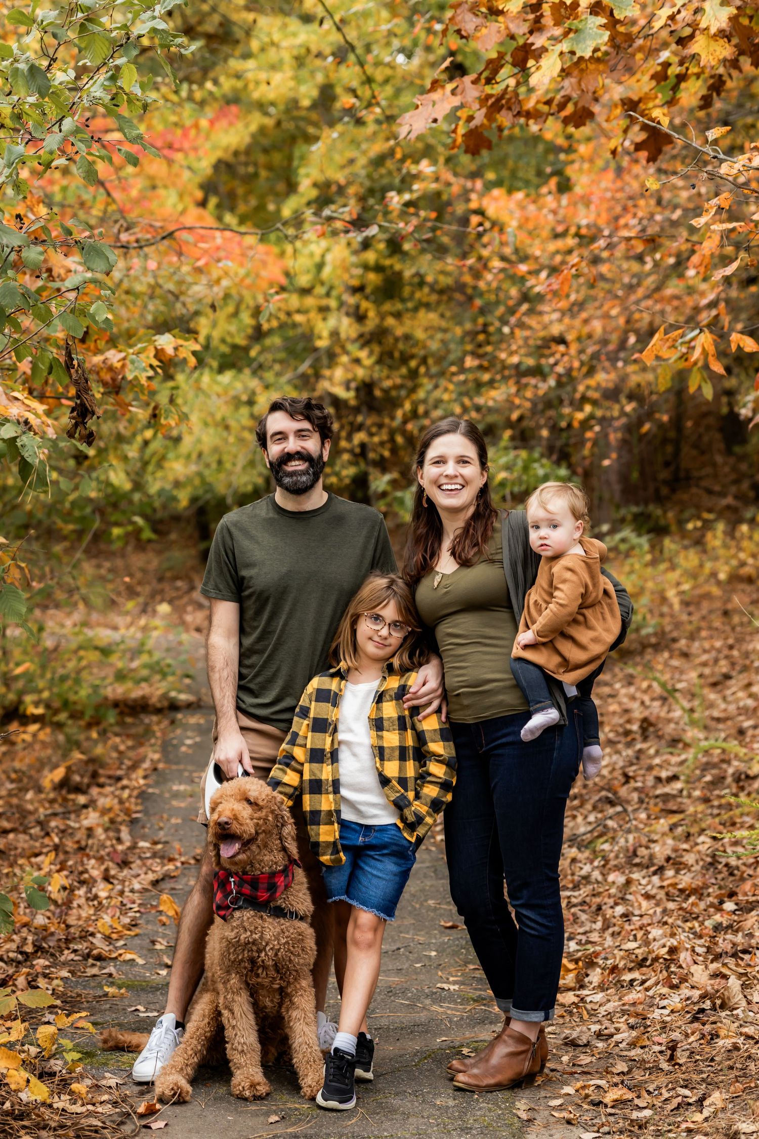 Family of four with a golden doodle posing on a leaf-covered path surrounded by vibrant autumn foliage.