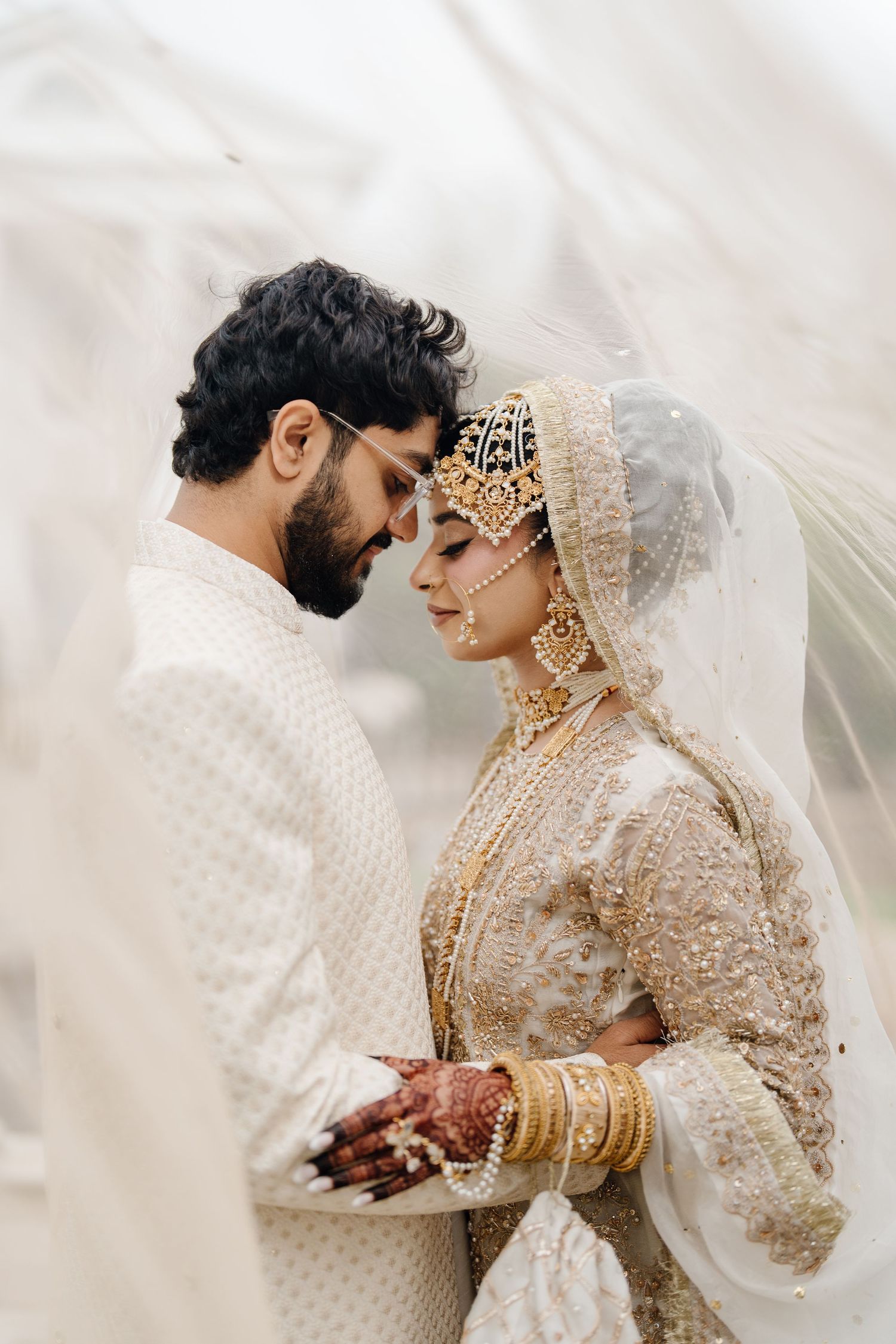 Indian bride in gold and red bridal lehenga seated before white floral arch backdrop with cascading blooms.