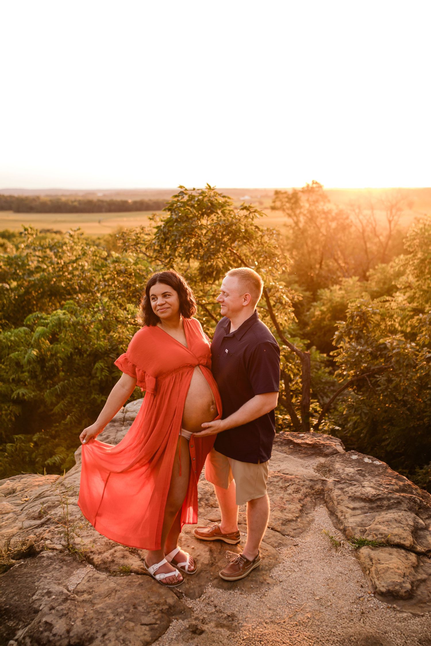 Pregnant couple posing on rocky overlook at golden sunset, woman in flowing red dress with partner touching her baby bump.