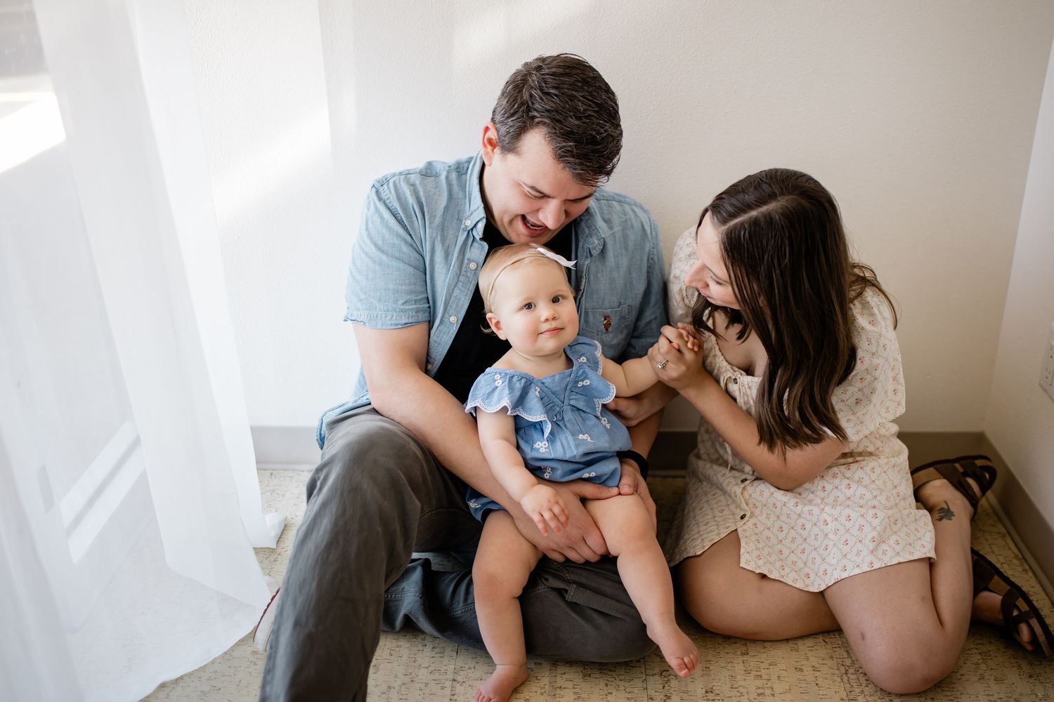 A happy young family with a baby girl in a blue romper sits together on a bright, airy floor in a warm moment.