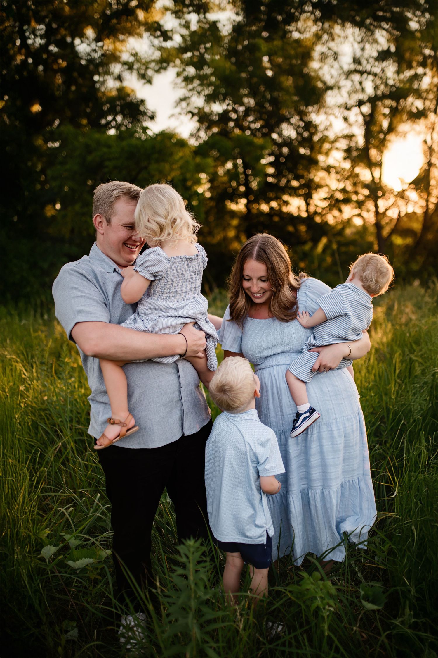 Family of five posing in a sunlit green meadow at golden hour, parents holding toddlers in matching blue outfits.