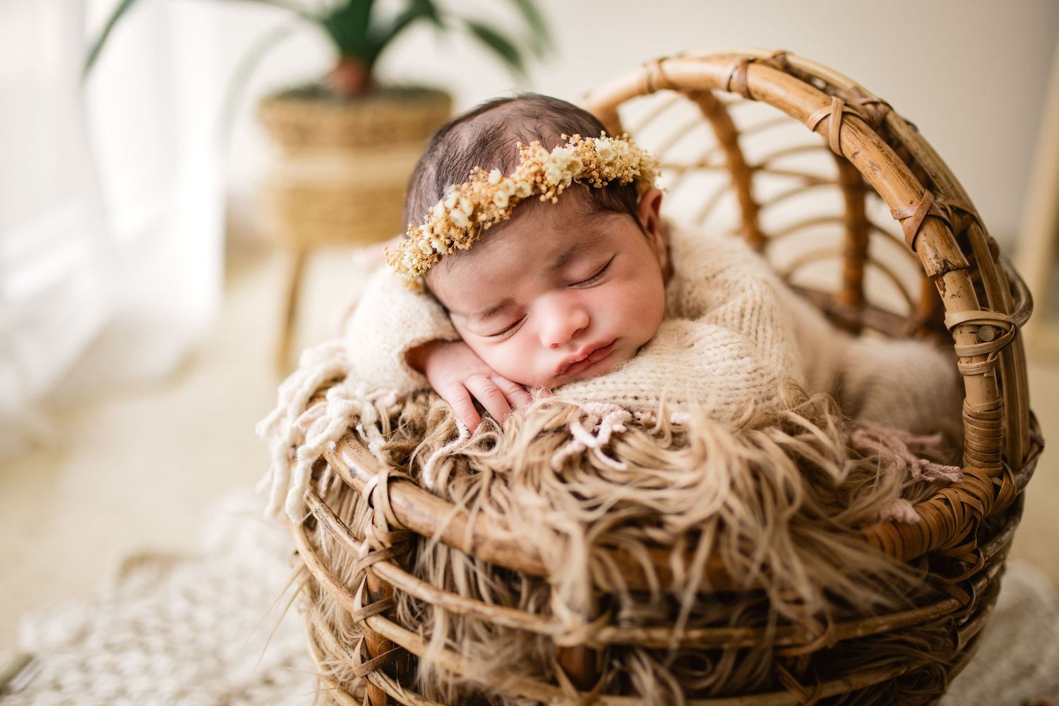 Sleeping newborn baby in a wicker basket with floral headband on fluffy fur blanket in soft natural light.