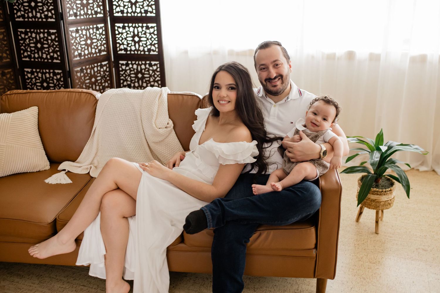 Happy family of three posing on a brown leather sofa, with a woman in white dress, man, and baby.