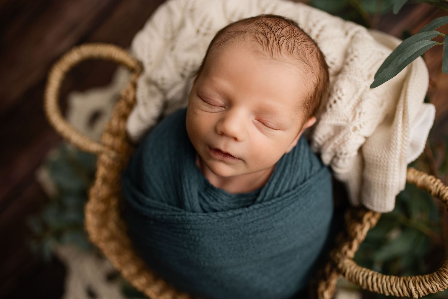 Newborn baby swaddled in teal wrap sleeping peacefully inside a wicker basket with green foliage backdrop.