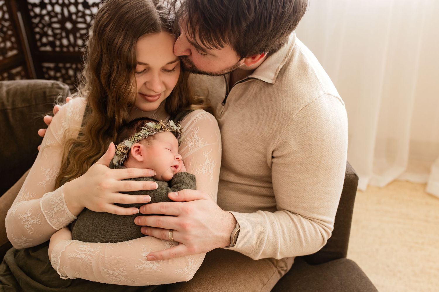 A couple tenderly holds their sleeping newborn baby, the father kissing the mother's head in a warm studio portrait.
