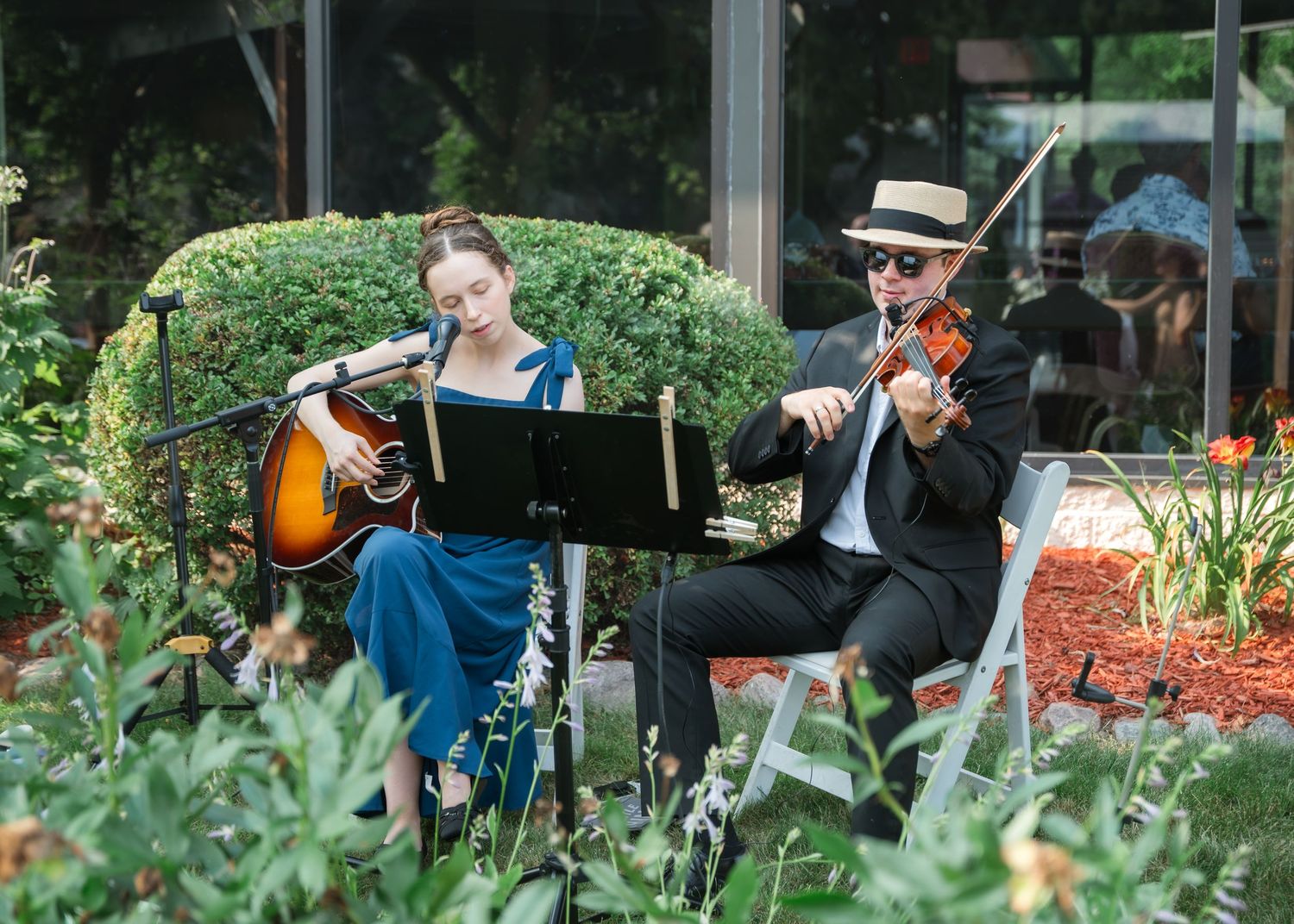 A woman playing guitar and a man playing violin perform outdoors amid lush greenery at an elegant garden event.