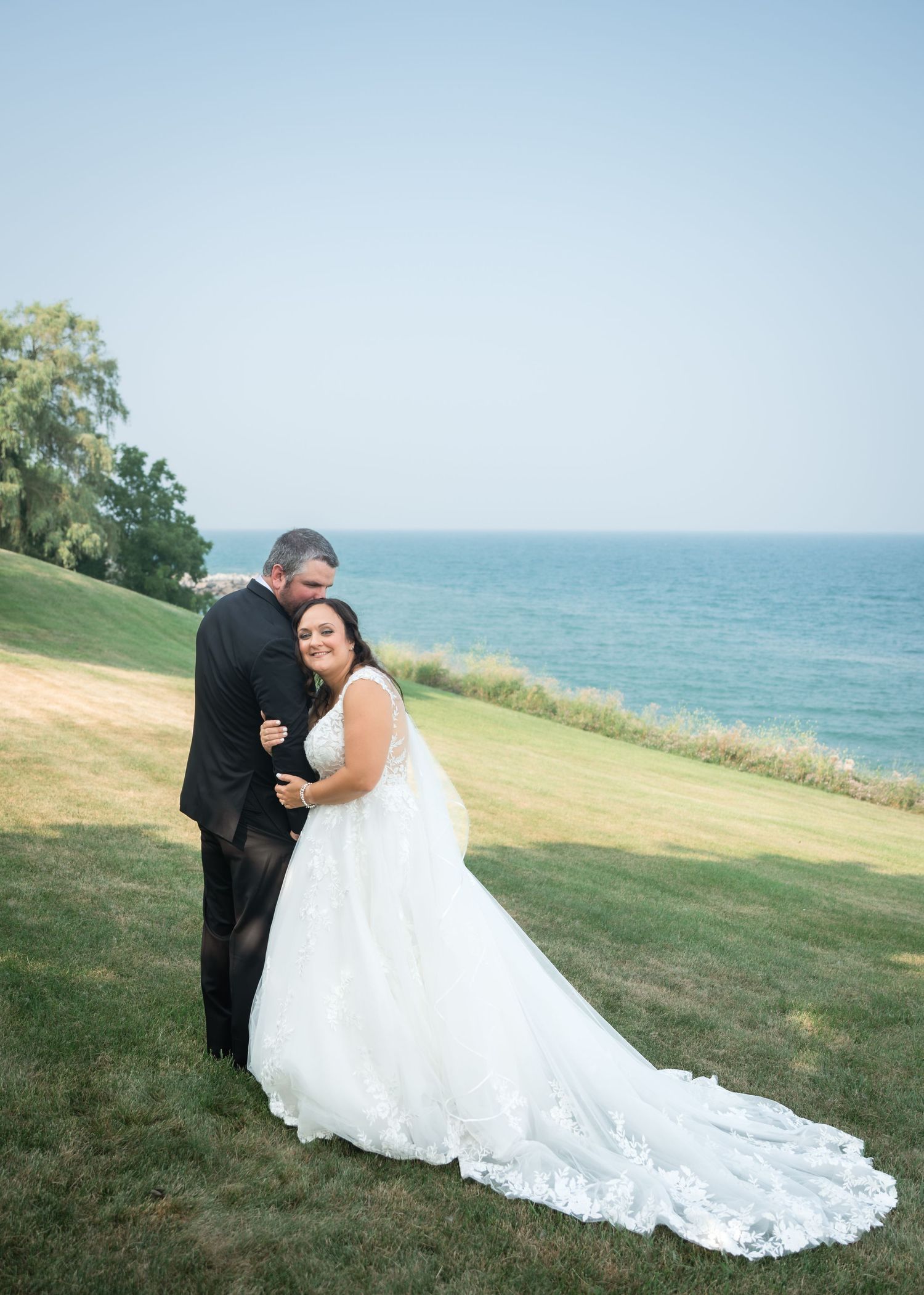 Bride in white lace gown and groom in black tux embrace on green lawn overlooking serene blue ocean.