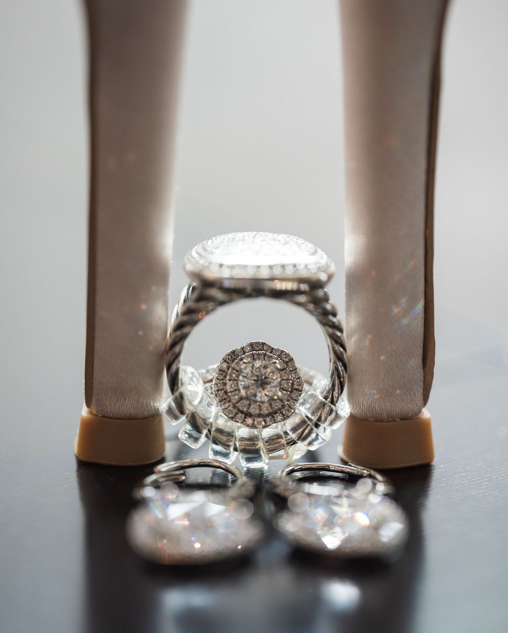 Diamond rings and jewelry displayed between white high heel shoes on a reflective surface.