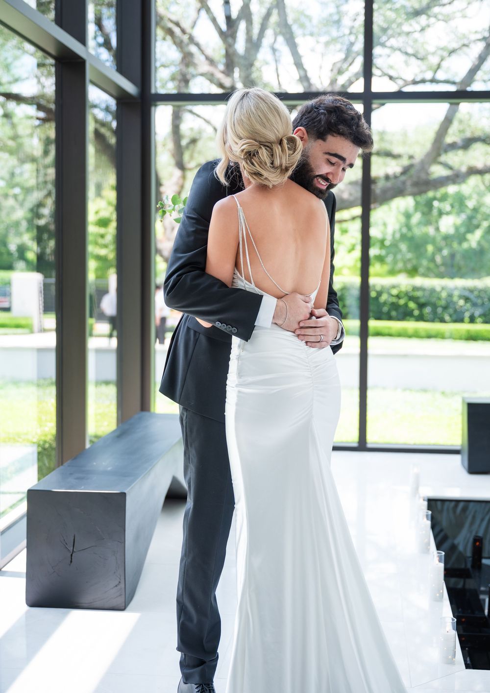 A bride in a backless white gown embraces her groom in a black suit near large windows with garden views.