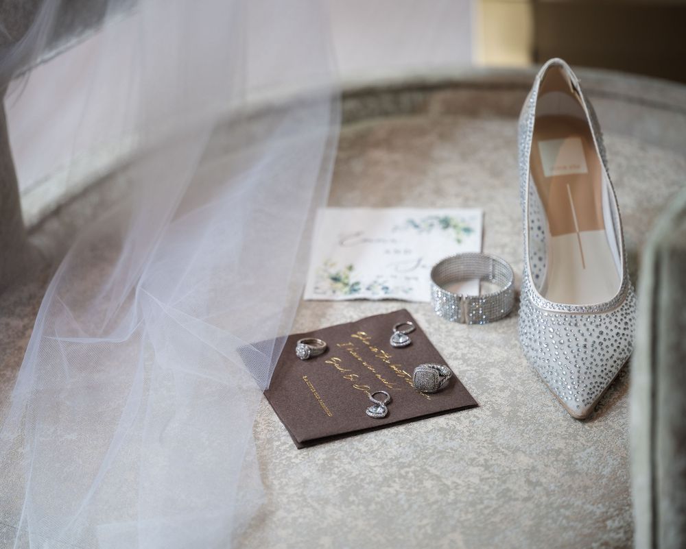 Elegant bridal accessories including crystal heels, veil, diamond rings, bracelet, and wedding invitation on marble surface.