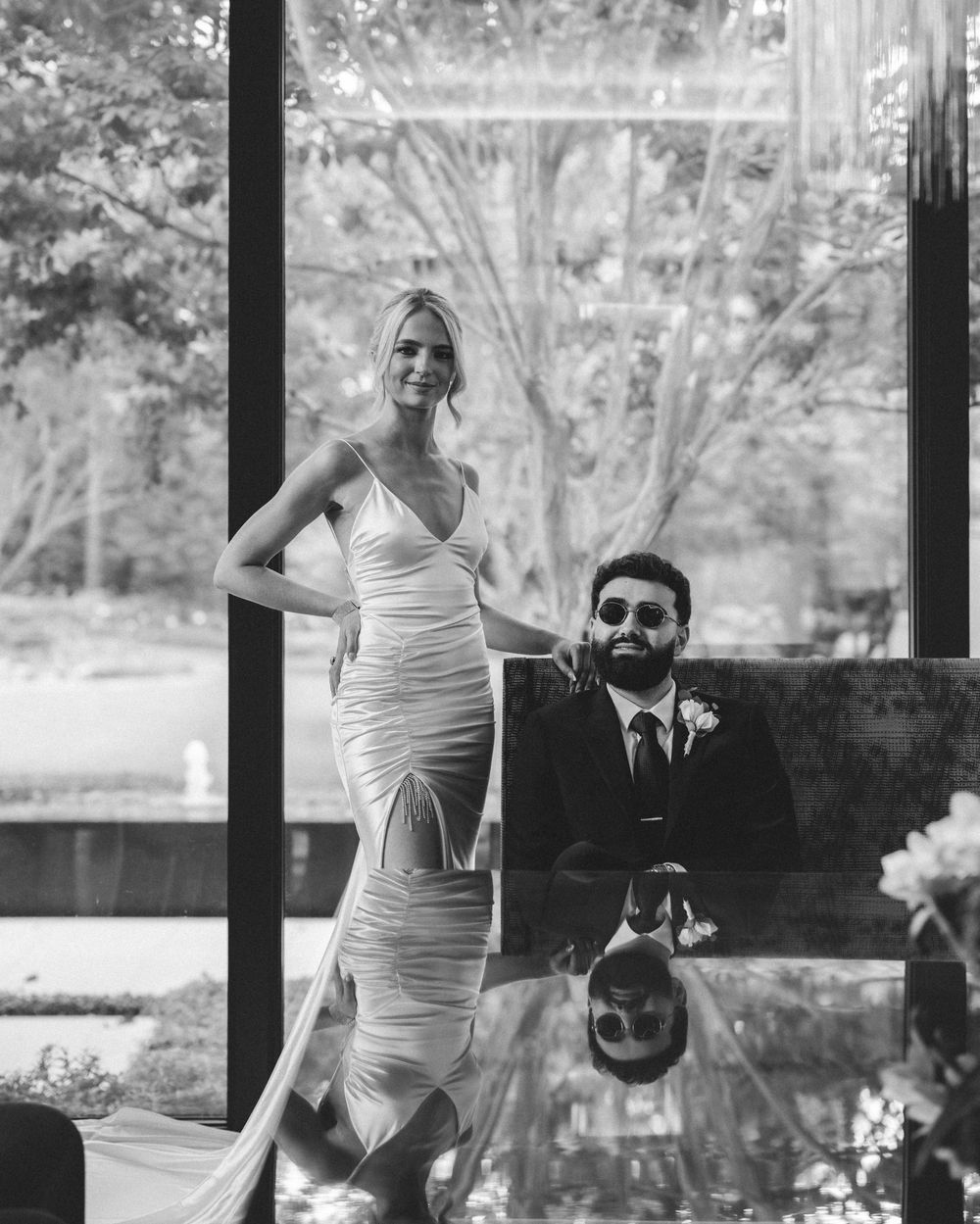 Black and white wedding photo of bride in elegant gown and groom in suit posing by reflective table in glass venue.