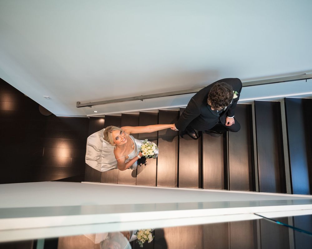 Bride and groom photographed from above on modern staircase, bride holding bouquet in elegant wedding attire.