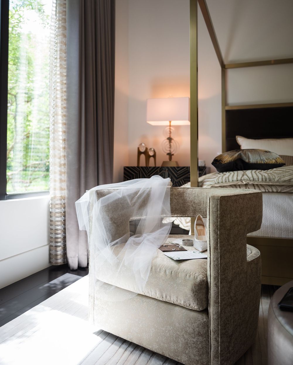 Elegant bedroom with a velvet armchair, four-poster bed, bedside lamp, and sheer curtains framing a window with green views.