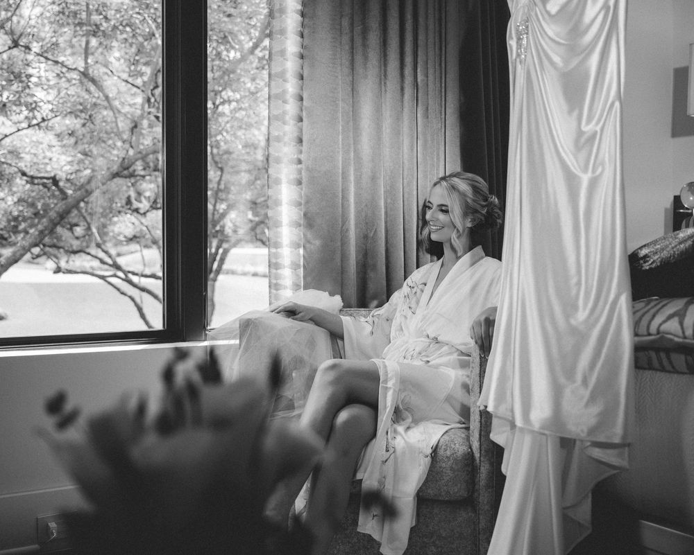 Bride in white robe smiling in chair by window, wedding dress hanging nearby, black and white photo.