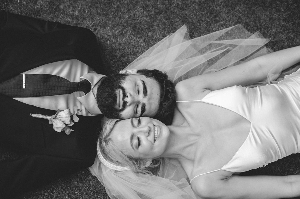 Black and white photo of smiling bride and groom lying on grass, holding hands on their wedding day.