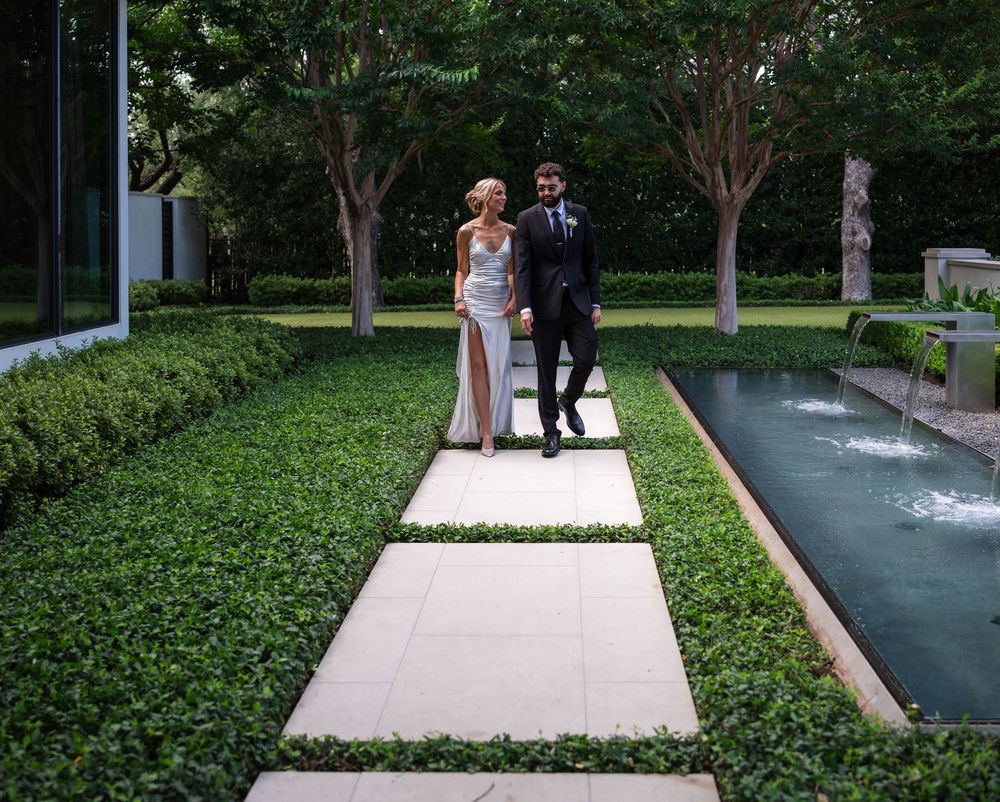 A bride and groom walk hand in hand along a stone path surrounded by lush green hedges and a reflecting pool.