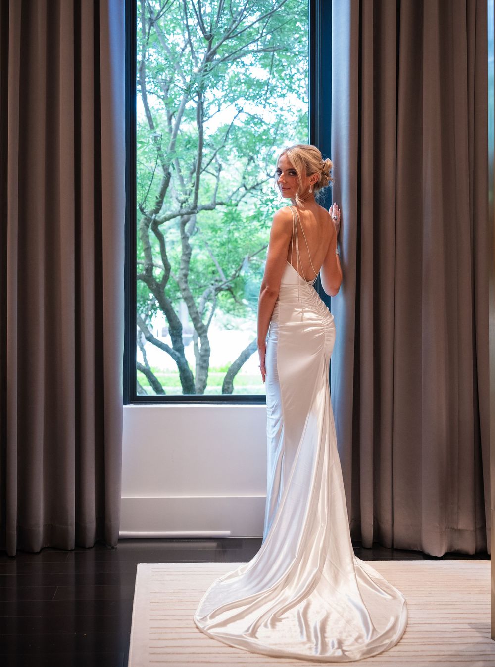 Bride in elegant backless white satin wedding gown with train stands by large window with flowing curtains.