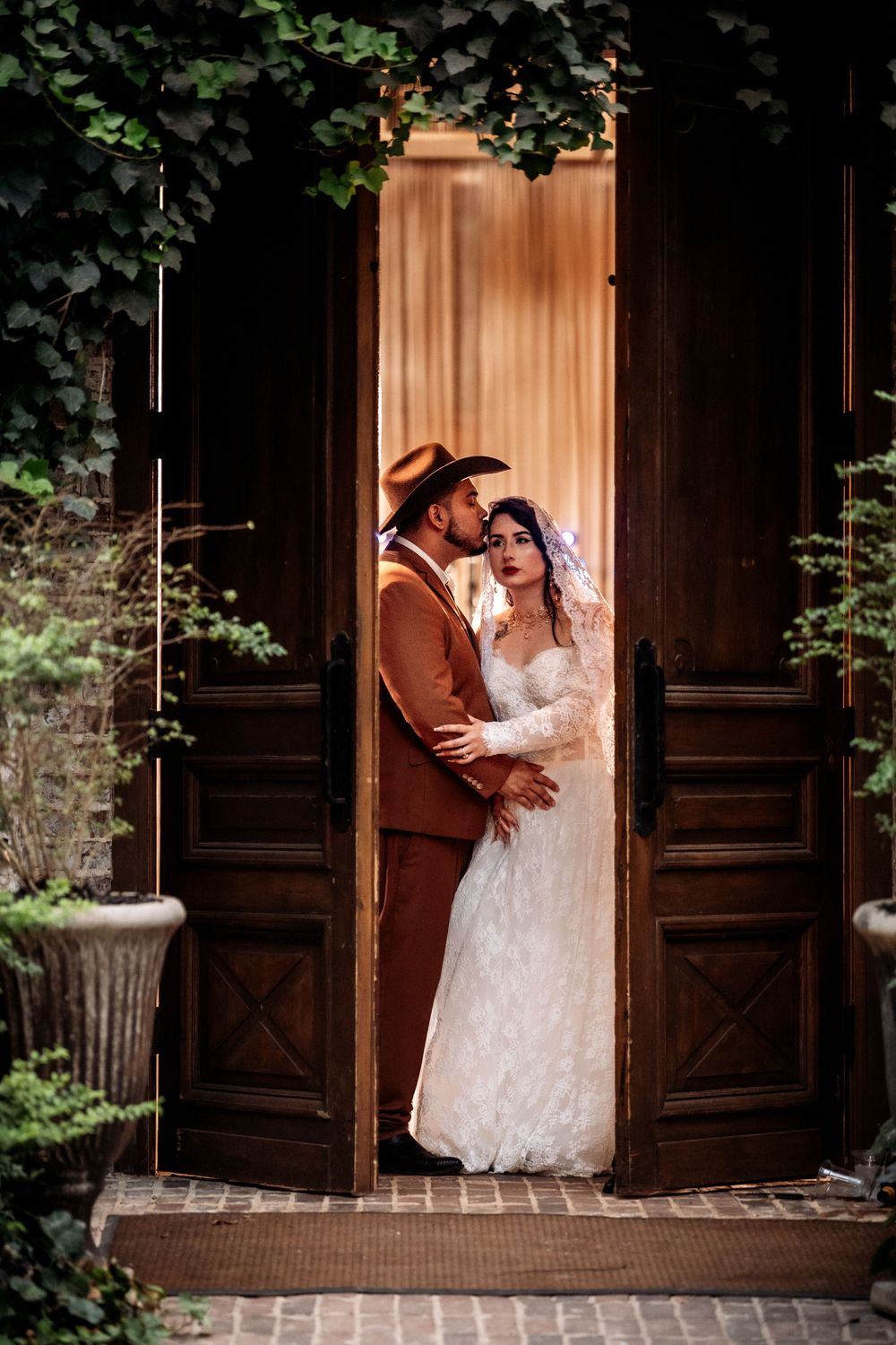A romantic couple in wedding attire shares an intimate moment in front of wooden double doors surrounded by greenery.