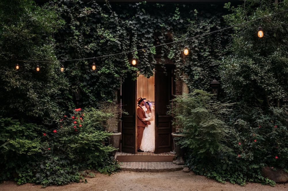 String lights illuminate a lush garden pathway leading to a rustic wooden doorway at dusk.